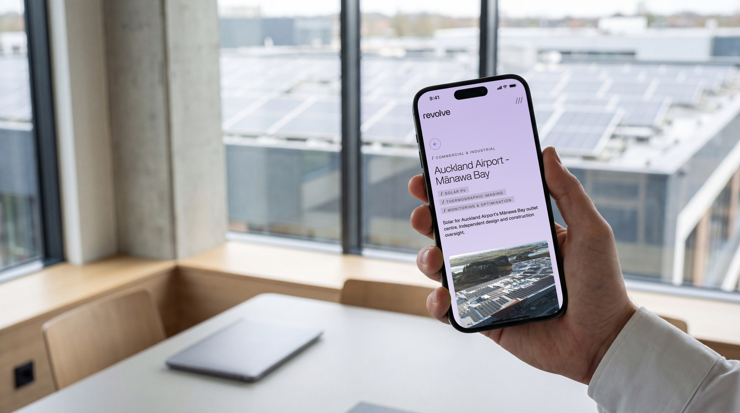 Revolve Energy's Auckland Airport project page on a phone, held in an office overlooking solar panels